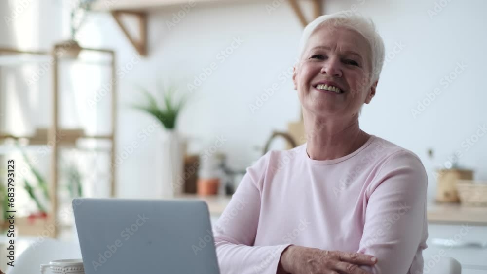 Senior adult Caucasian woman gazes into the camera with a sense of purpose, her hands resting on the keyboard of her laptop on the kitchen table. Her eyes convey wisdom and experience.