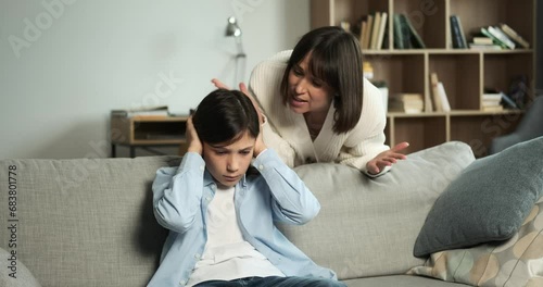 Furious mother reprimands son, who sits on the couch, receiving scolding. The tension in the room is palpable, showcasing a difficult moment of parental discipline and family dynamics. photo