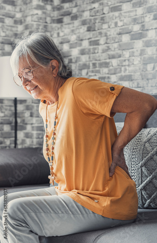 Unhappy mature woman touching back, feeling pain, sitting on couch in living room, unhealthy middle aged senior female suffering from backache, spinal problem, rubbing stiff muscles or kidney. photo