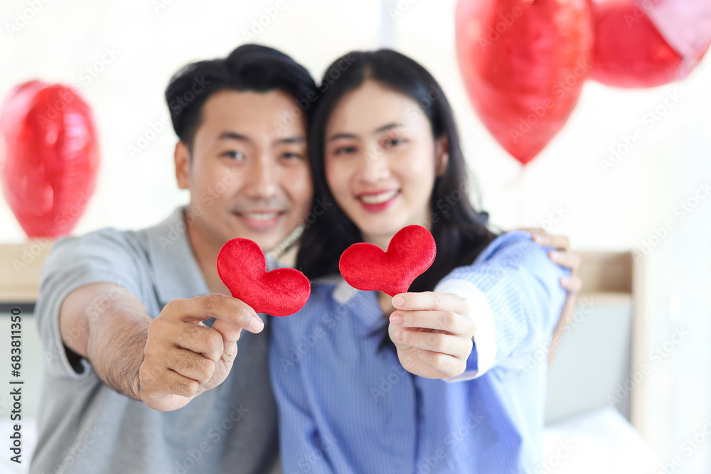 Two red hearts on hands of happy Asian couple who sitting on bed in beautifully decorated bedroom with balloons, romantic lover celebrating anniversary on Valentine Day together, love celebration. Two red hearts on hands of happy Asian couple who sitting on bed in beautifully decorated bedroom with balloons, romantic lover celebrating anniversary on Valentine Day together, love celebration.