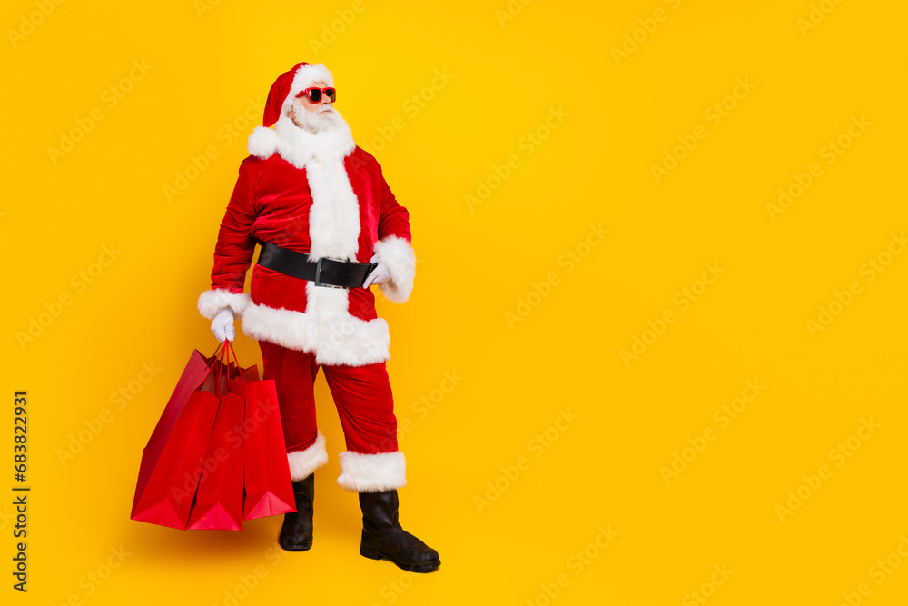 Full length photo of santa red costume sunglass touch belt look empty space to buy shopping mall bags isolated on yellow color background Full length photo of santa red costume sunglass touch belt look empty space to buy shopping mall bags isolated on yellow color background