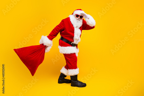 Full body profile portrait of eccentric santa claus arm touch forehead hold new year gifts bag empty space isolated on yellow color background