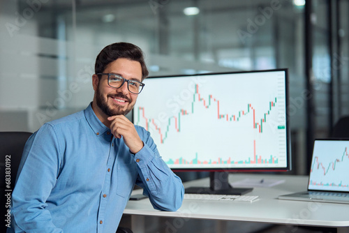Young confident latin business man trader crypto investor broker looking at camera, portrait. Digital trade stock exchange charts on computer screen, investing in financial markets concept. photo
