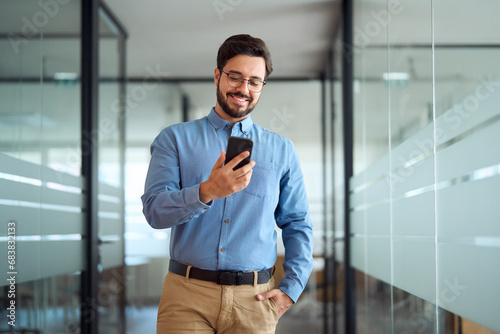 Smiling busy professional latin business man walking in office hallway holding mobile cellphone. Young happy businessman employee using smartphone looking at cell phone tech standing at work. photo