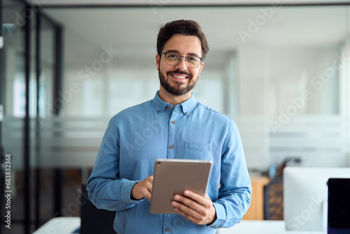 Smiling busy young latin business man manager using tablet computer, happy hispanic businessman executive looking at camera holding tab working standing in office. Portrait. photo