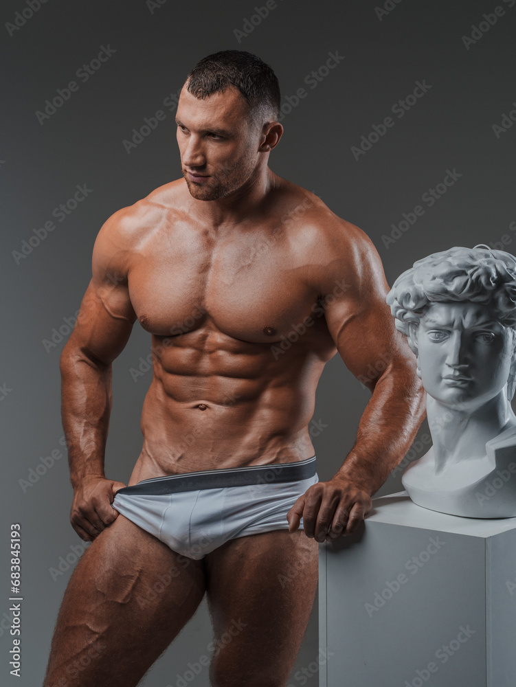 Fototapeta premium Handsome man exuding a rugged charm confidently showcasing his immaculate muscular physique while standing beside ancient Greek statues against a gray backdrop