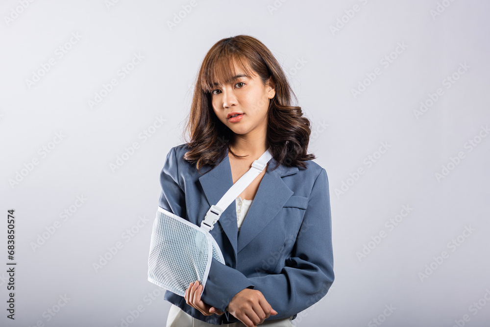 Asian businesswoman, happily coping with a painful broken arm, wears a splint for treatment. Sling support in the studio, isolated on white with copy space. Her injury is clear. Asian businesswoman, happily coping with a painful broken arm, wears a splint for treatment. Sling support in the studio, isolated on white with copy space. Her injury is clear.