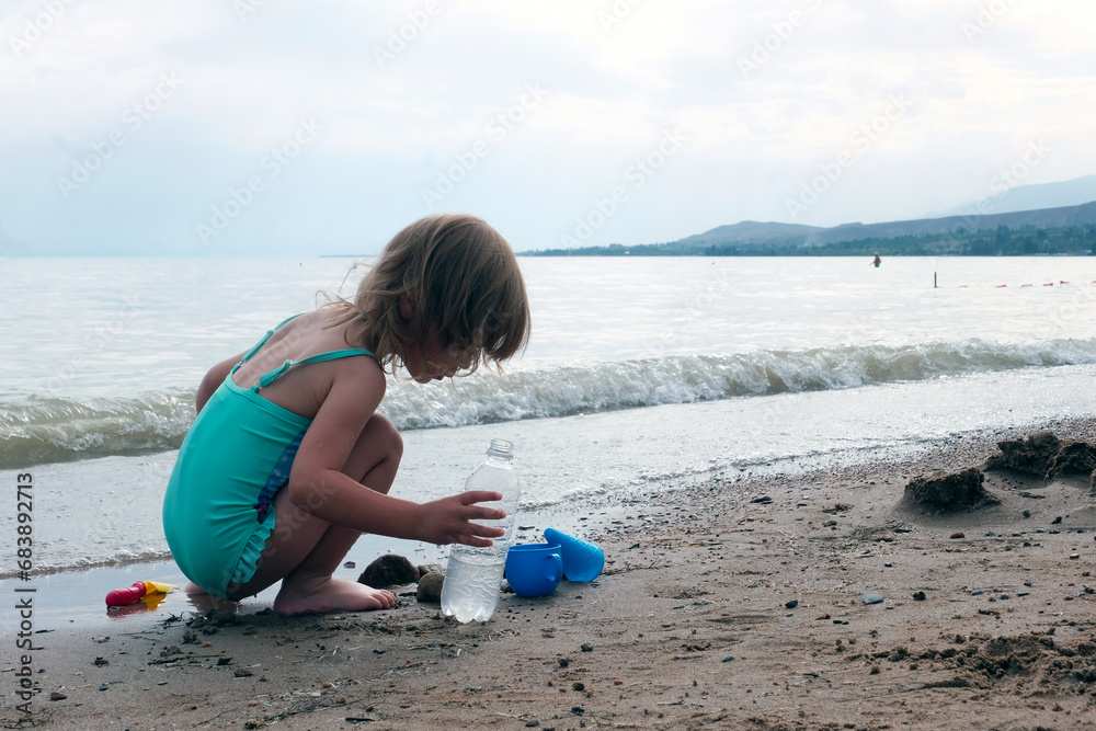 A little fair-haired girl in a green swimsuit plays with toys on the seashore. Concept of safe water games, healthy lifestyle, tourism. A little fair-haired girl in a green swimsuit plays with toys on the seashore. Concept of safe water games, healthy lifestyle, tourism.