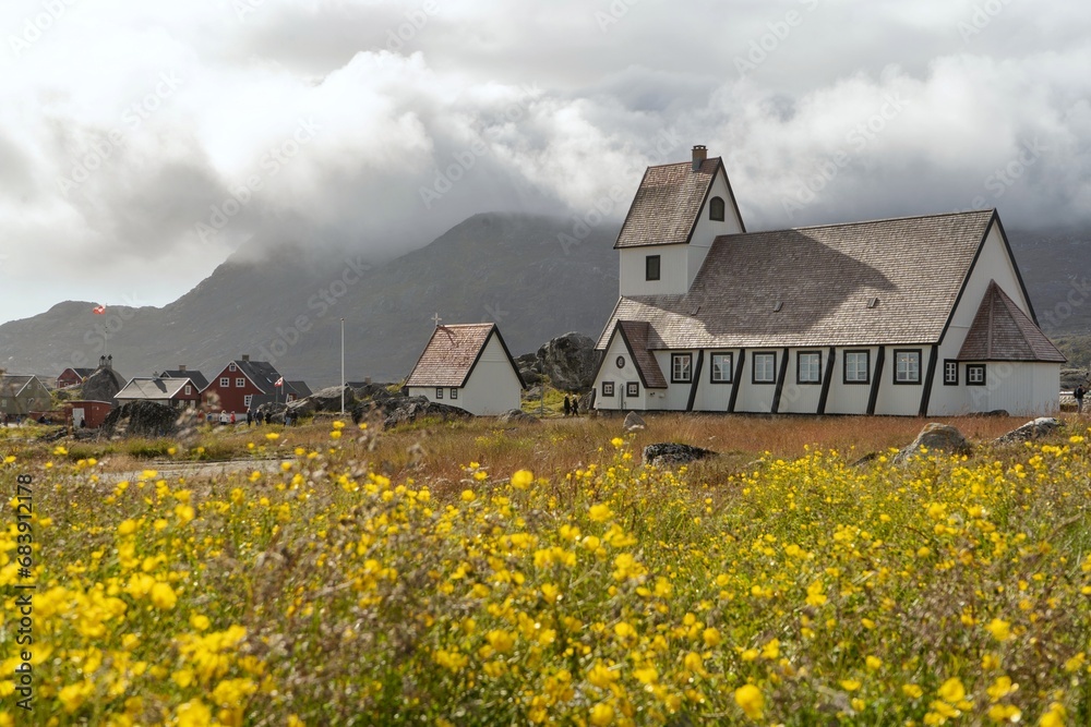 Landscape with a wooden cathedral in mountains in Greenland, Nanortalik, with field of flowers, with a flags, with clouds, during summer Landscape with a wooden cathedral in mountains in Greenland, Nanortalik, with field of flowers, with a flags, with clouds, during summer