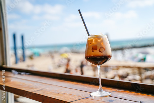 A glass with an cold coffee alcoholic drink and ice in a beach restaurant with a straw