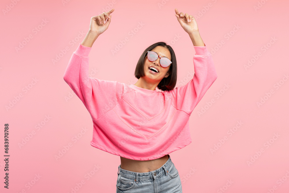 Portrait of happy attractive Asian woman wearing sunglasses hands up dancing looking at camera
