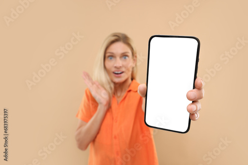 Surprised woman holding smartphone with blank screen on beige background, selective focus