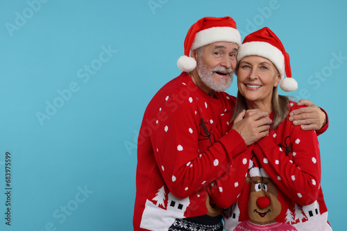 Senior couple in Christmas sweaters and Santa hats on light blue background. Space for text