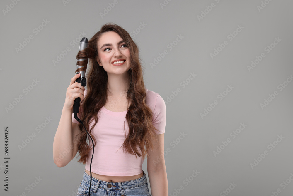 Beautiful young woman using curling hair iron on grey background, space for text Beautiful young woman using curling hair iron on grey background, space for text