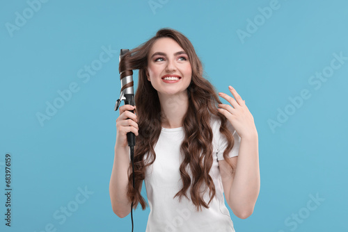 Beautiful young woman using curling hair iron on light blue background