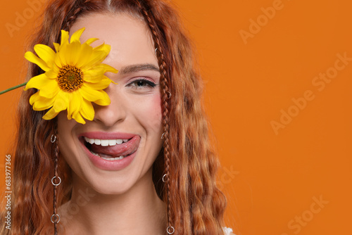 Beautiful young hippie woman covering eye with sunflower and showing her tongue on orange background, closeup. Space for text