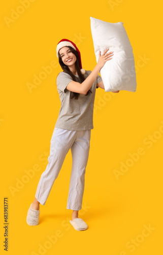 Beautiful young woman in pajamas and Santa hat with soft pillow on yellow background © Pixel-Shot