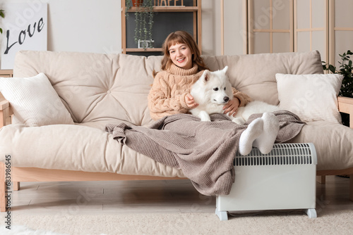 Young woman with blanket and Samoyed dog on sofa warming near radiator at home