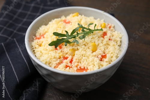 Tasty couscous with pepper, corn and arugula in bowl on wooden table, closeup