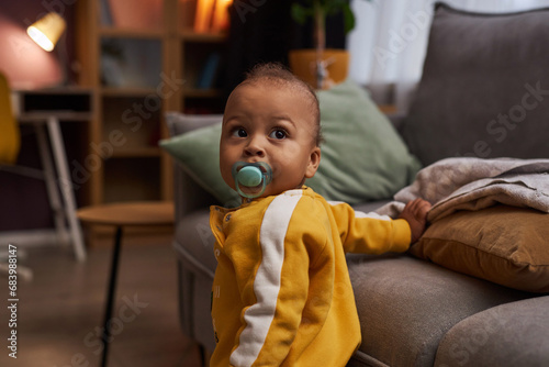 Portrait of cute baby boy with binky taking first steps in cozy living room, copy space photo