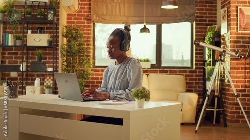 Freelancer enjoys working at workstation while listening to music and performing internet activities. At sunset, african american woman is multitasking, online career in blogging.