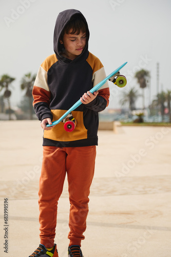 Caucasian teenage boy dressed in stylish sportswear, holding skateboard and looking down, standing at outdoor skatepark
