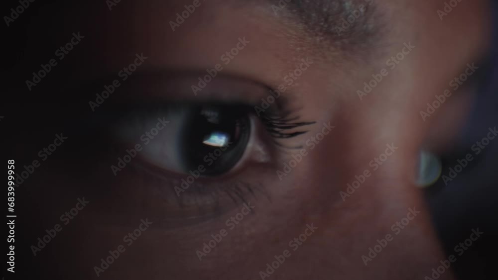 Close-up of young black woman eyes looking at device screen, working with computer laptop. Monitor light is reflected in her face. Female freelancer on Internet at dark night in living room.