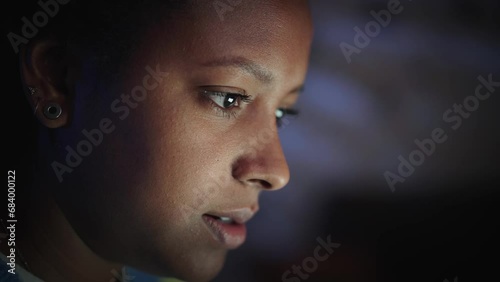 Side close-up face beautiful young American black woman looking at screen working with laptop indoor. Female freelancer typing on computer at night in home room. Concept studying gen z. photo