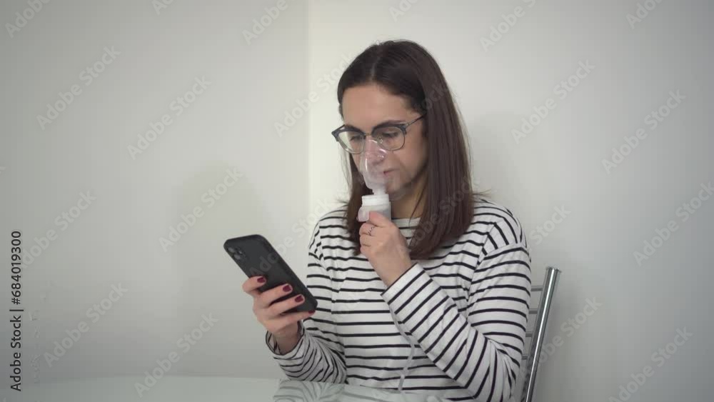 A young woman breathes through an inhaler mask with a phone in her hands. A girl in glasses with an oxygen mask is being treated for a respiratory infection and chatting on her smartphone. A young woman breathes through an inhaler mask with a phone in her hands. A girl in glasses with an oxygen mask is being treated for a respiratory infection and chatting on her smartphone.
