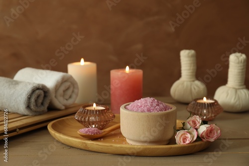 Bowl of pink sea salt, roses, burning candles, herbal massage bags and towels on wooden table