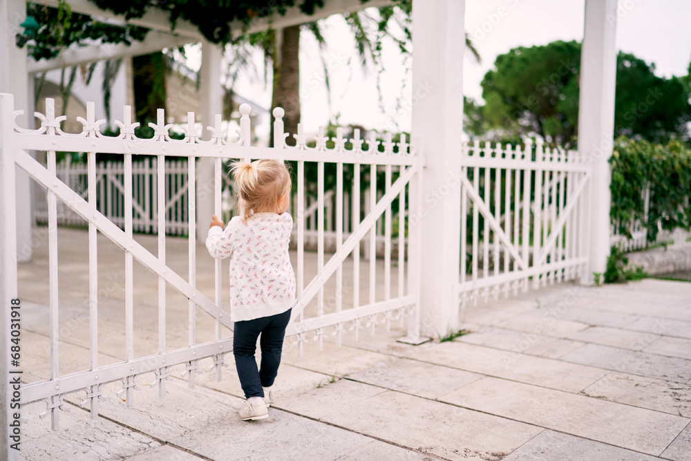 Little girl stands holding on to the bars of a metal fence in the park and looks into the distance. Back view
