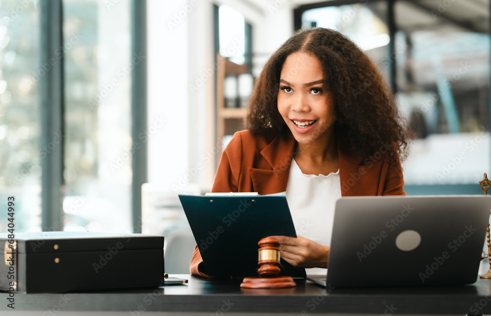 Attractive smiling African American female lawyer reads a document in an office, with a laptop, gavel, and scales of justice nearby, legal, legislation. Attractive smiling African American female lawyer reads a document in an office, with a laptop, gavel, and scales of justice nearby, legal, legislation.
