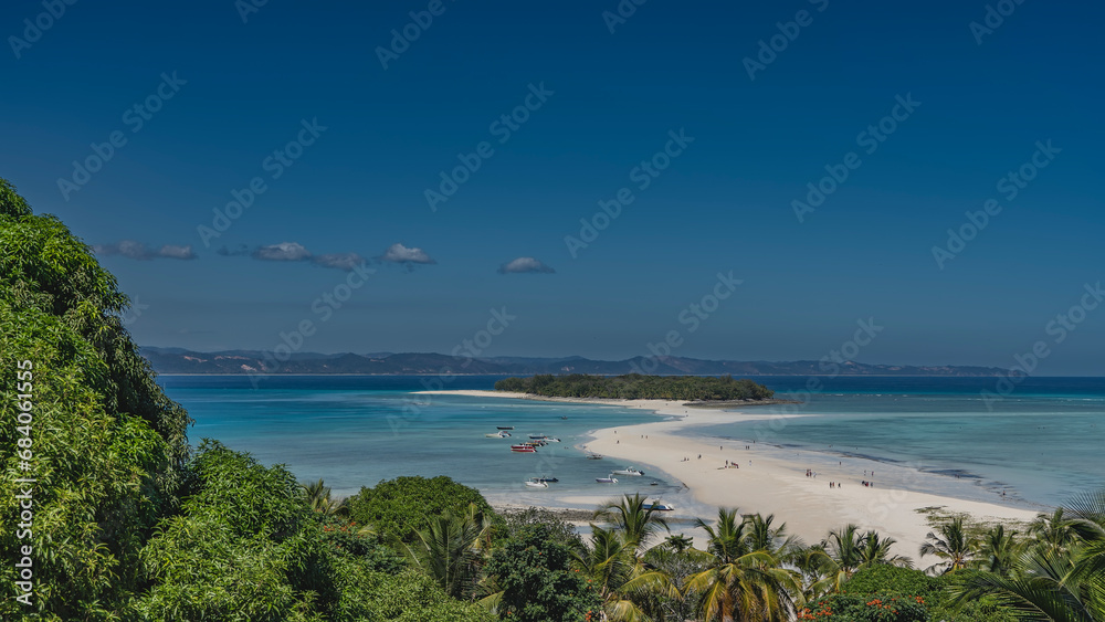 A beautiful tropical island and a winding sand spit are visible in the turquoise ocean. Tiny silhouettes of people on the beach. Boats at the water's edge. In the foreground is lush green vegetation.