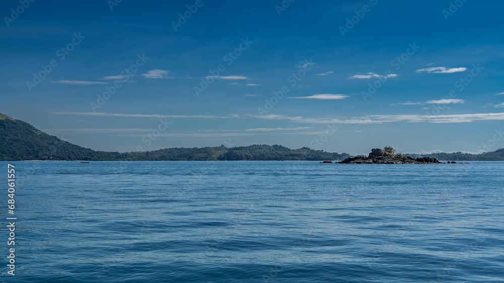 A small rocky island in the blue ocean. Ripples on the water. A mountain range in the distance against the background of azure sky and clouds. Madagascar.