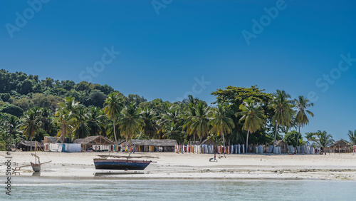 Malagasy village on the ocean. Among the palm trees- houses with thatched roofs, drying laundry. Wooden fishing boats at the water's edge. A hill with tropical vegetation against a clear blue sky. 