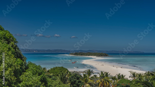 A beautiful tropical island and a winding sand spit are visible in the turquoise ocean. Tiny silhouettes of people on the beach. Boats at the water's edge. In the foreground is lush green vegetation. © Вера 