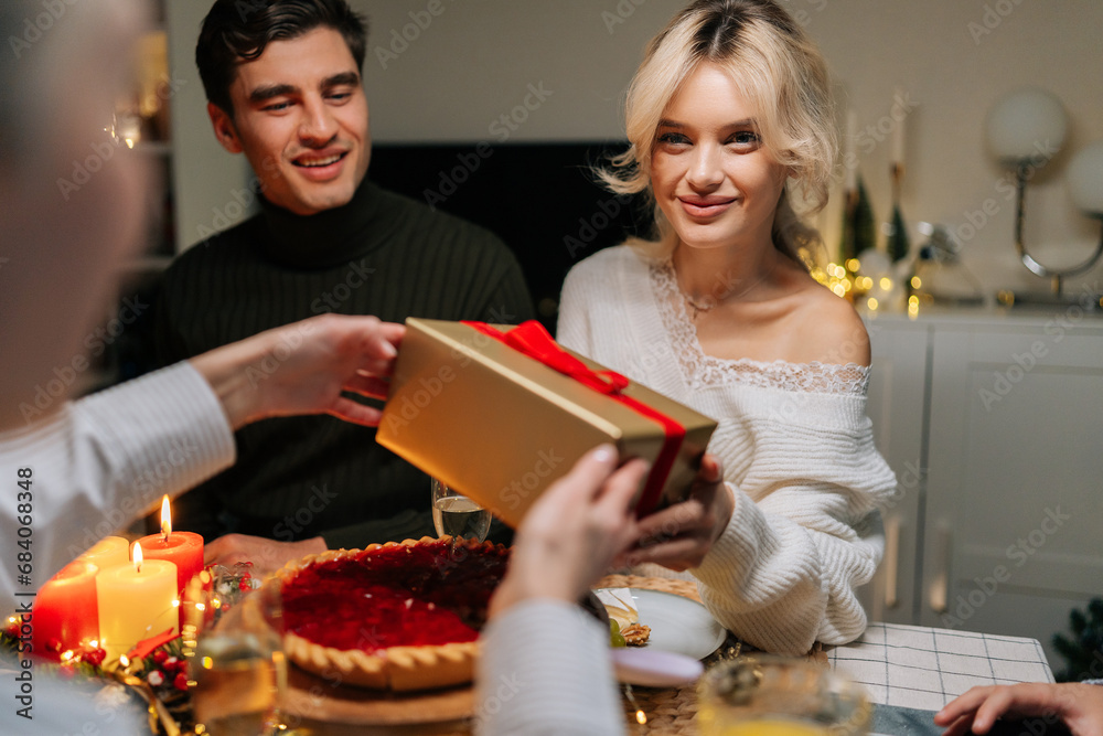 Front view of happy young woman receiving gift from loving mother-in-law sitting with family at dinner feast table at home on Christmas Eve. Blonde female giving present to beloved daughter on holiday