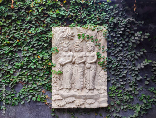 carvings of 3 traditional women on marble with a background of plants creeping on the wall