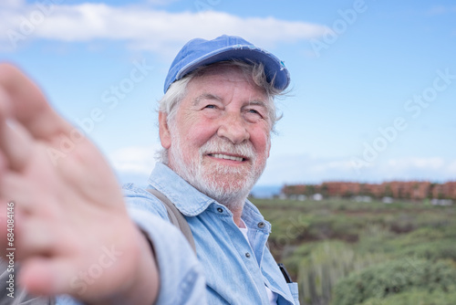 Portrait of senior happy man with hat walking in countryside footpath looking at camera. Elderly bearded grandparent smiling enjoying healthy lifestyle and nature.