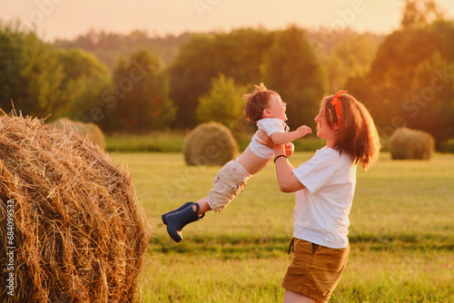 Mother holding her child, standing on a field with hay stacks amidst the setting sun, while the baby laughs with joy. Portrait of a happy kid two years old and a woman thirty five years old photo