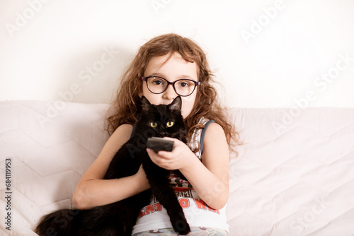 Cute curly little girl kid child in a eyeglasses watching TV with kitty cat best friend at home with remote control in a hands photo