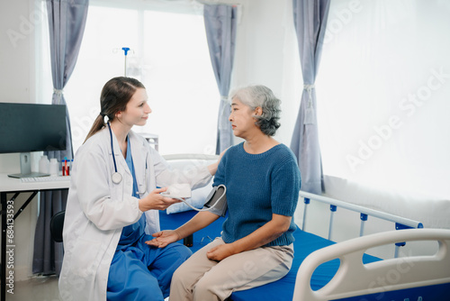 Doctor in white suit take notes while discussing and Asian elderly, woman patient who lying on bed with receiving saline solution in hospital or clinic.