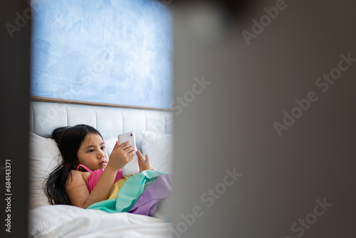 asian child or kid girl sleep lying pillow on bed for playing smartphone or addicted looking mobile phone to hyperactive ADHD and parents secretly watch children or baby behind ajar door in bedroom
