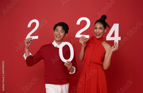 Happy asian couple in red casual attire showing number 2024 greeting and bless happy new year with smiles isolated on red background.