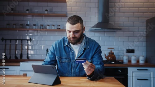 Young adult man sitting against the kitchen counter entering credit card number on tablet device for makes secure easy distant electronic payment at home photo