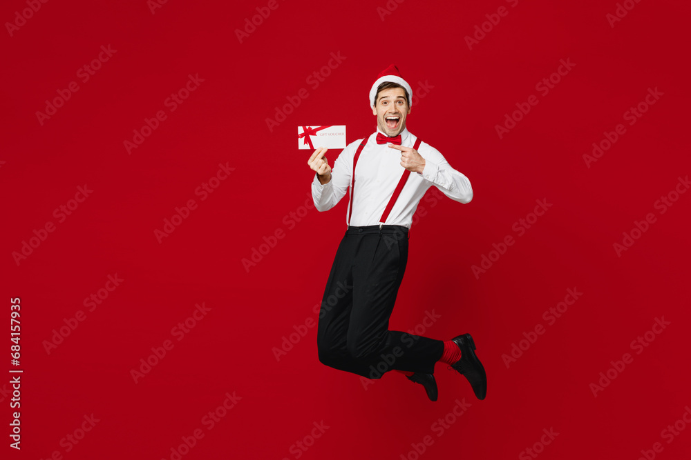 Full body merry young man wear white shirt Santa hat posing jump high point on gift coupon voucher card for store isolated on plain red background Happy New Year Christmas celebration holiday concept Full body merry young man wear white shirt Santa hat posing jump high point on gift coupon voucher card for store isolated on plain red background Happy New Year Christmas celebration holiday concept