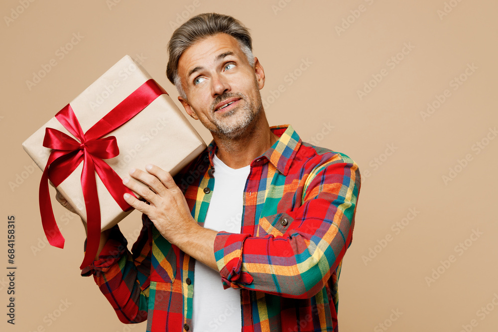 Adult man wear red shirt white t-shirt casual clothes hold in hand present box with gift ribbon bow look aside isolated on plain pastel light beige color background studio portrait. Lifestyle concept. Adult man wear red shirt white t-shirt casual clothes hold in hand present box with gift ribbon bow look aside isolated on plain pastel light beige color background studio portrait. Lifestyle concept.