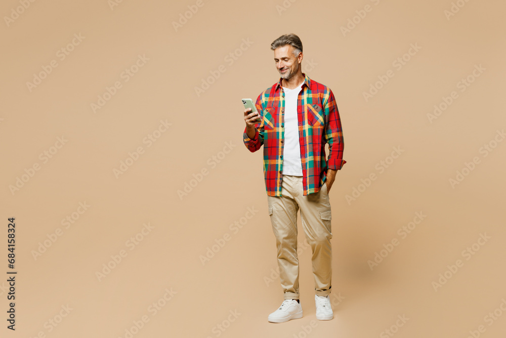 Full body adult man wear red shirt white t-shirt casual clothes hold in hand use mobile cell phone chat online isolated on plain pastel light beige color background studio portrait. Lifestyle concept. Full body adult man wear red shirt white t-shirt casual clothes hold in hand use mobile cell phone chat online isolated on plain pastel light beige color background studio portrait. Lifestyle concept.