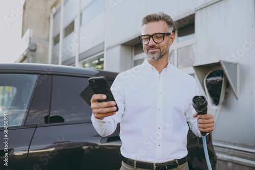 Adult confident man wearing shirt use mobile cell phone hold in hand electric car charger connect to electric car auto at charging station outdoor. Green energy solution for automobile fuel concept. photo