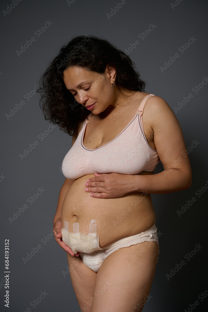Authentic woman, new mother touching her belly with a bandage hiding scars after c-section, few days after childbirth Authentic woman, new mother touching her belly with a bandage hiding scars after c-section, few days after childbirth